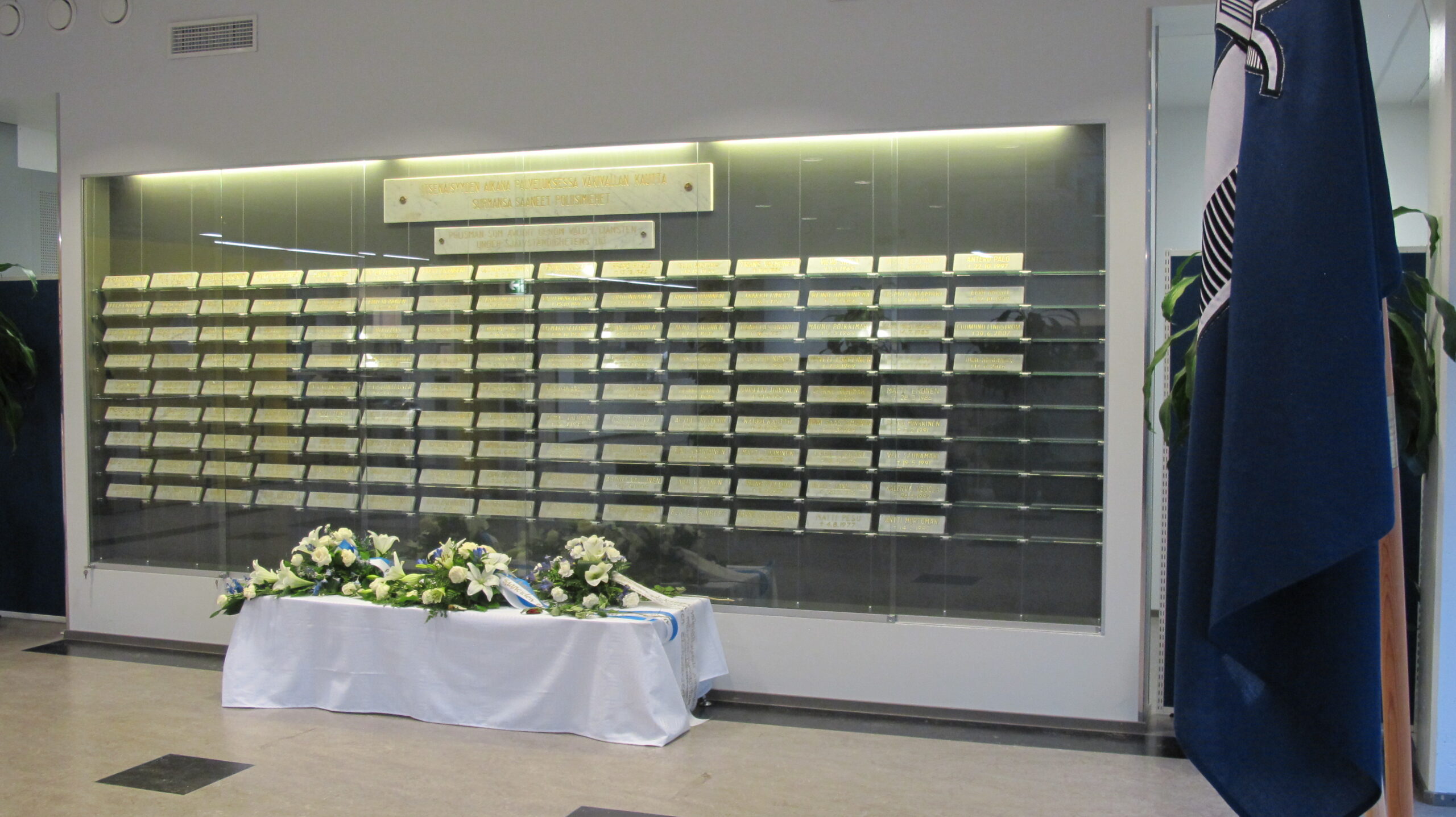 Lasivitriinissä useita vaakasuoria, suorakulmaisia muistolaattoja. Vitriinin yläpuolella on kaksi valaistu kylttiä. Vitriinin edessä on valkoisella liinalla peitetty pöytä, jonka päällä on kukka-asetelmia. Pöydän vieressä oikealla on tummansininen lippu.