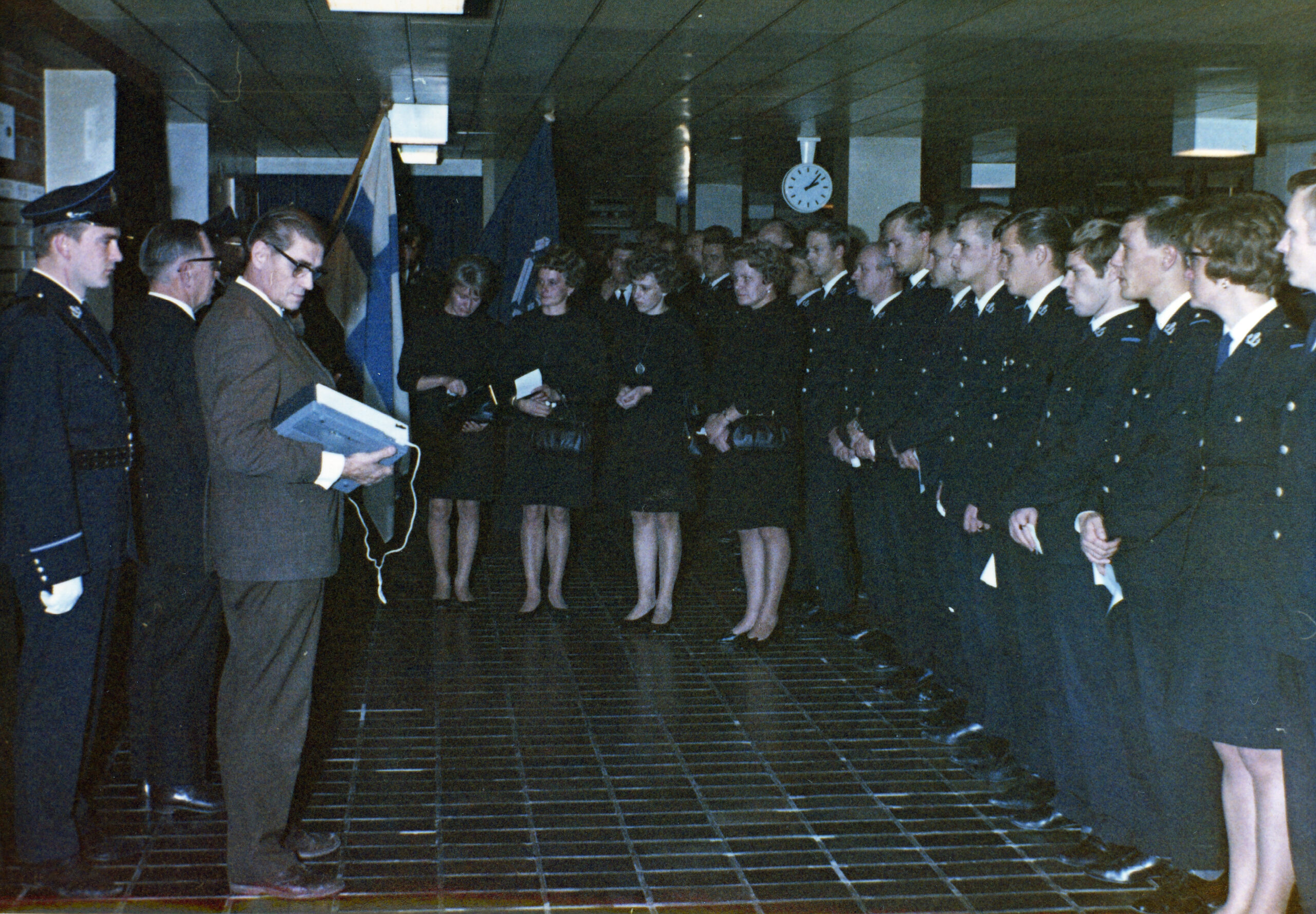 Värillinen kuva tilaisuudesta, jossa rivi tummiin univormuihin pukeutuneita ihmisiä seisoo. Edessä siviilipukuinen mies lukee papereita Suomen lipun vieressä.