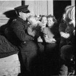 Mustavalkoinen kuva, jossa virkapukuinen poliisi pitää pientä lasta sylissään. Taka-alalla hymyileviä naisia.