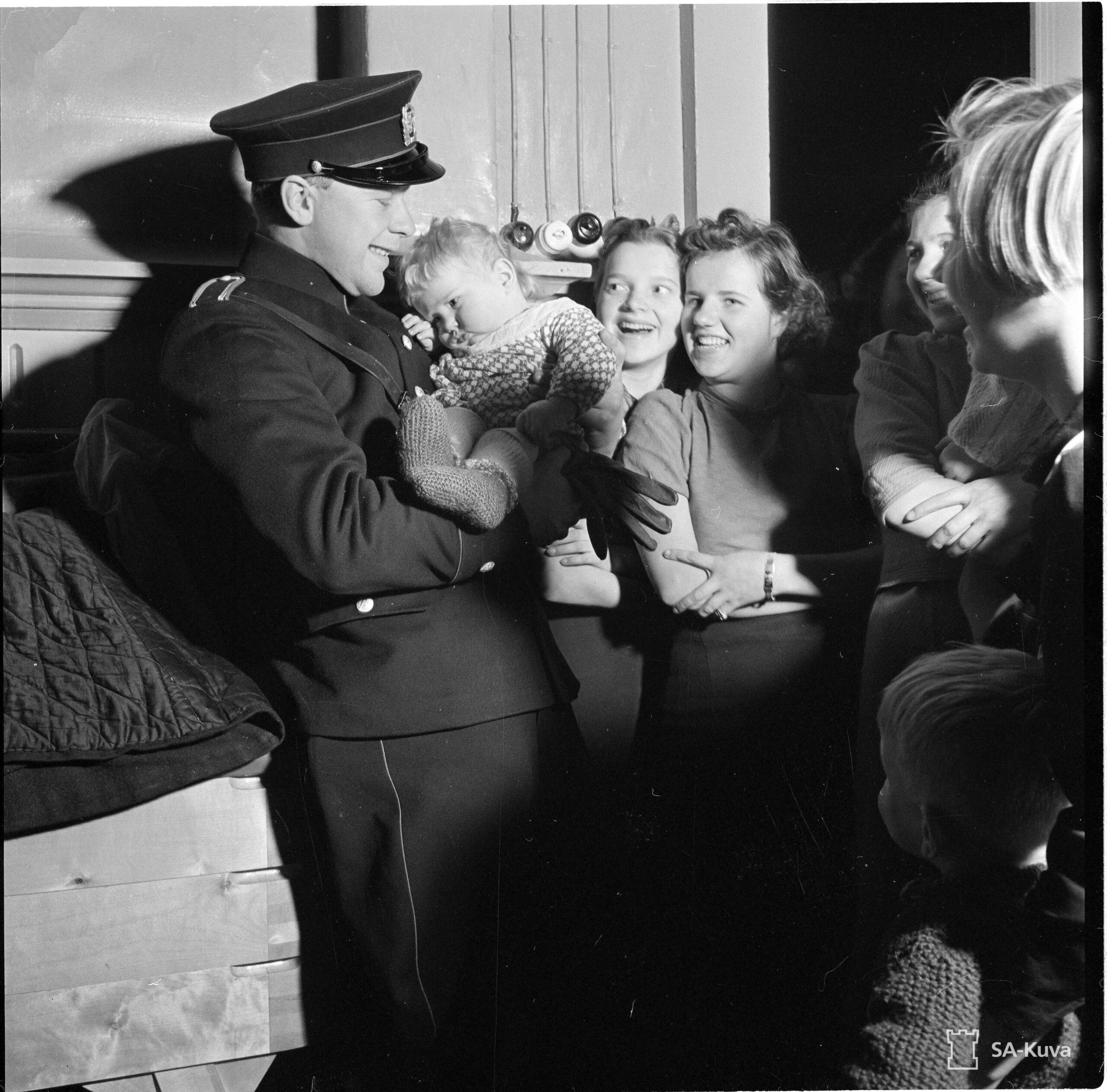 Mustavalkoinen kuva, jossa virkapukuinen poliisi pitää pientä lasta sylissään. Taka-alalla hymyileviä naisia.