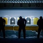 Värikuva, jossa talvella kolme henkilöä on valmistautumassa ampumaan maalitauluun ampumaradalla. Taulujen yläpuolella on numeroita.