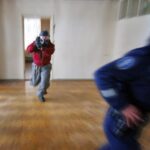 Värikuva, jossa sisätiloissa kaksi hahmoa juoksevat peräkkäin. Etualalla on poliisi, ja taustalla toinen henkilö juoksee punaiseen takkiin pukeutuneena ja miekka kädessä.