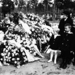 Mustavalkoinen kuva, jossa nainen ja kolme lasta seisovat lähekkäin haudalla, joka on peitetty seppeleillä ja kukilla.