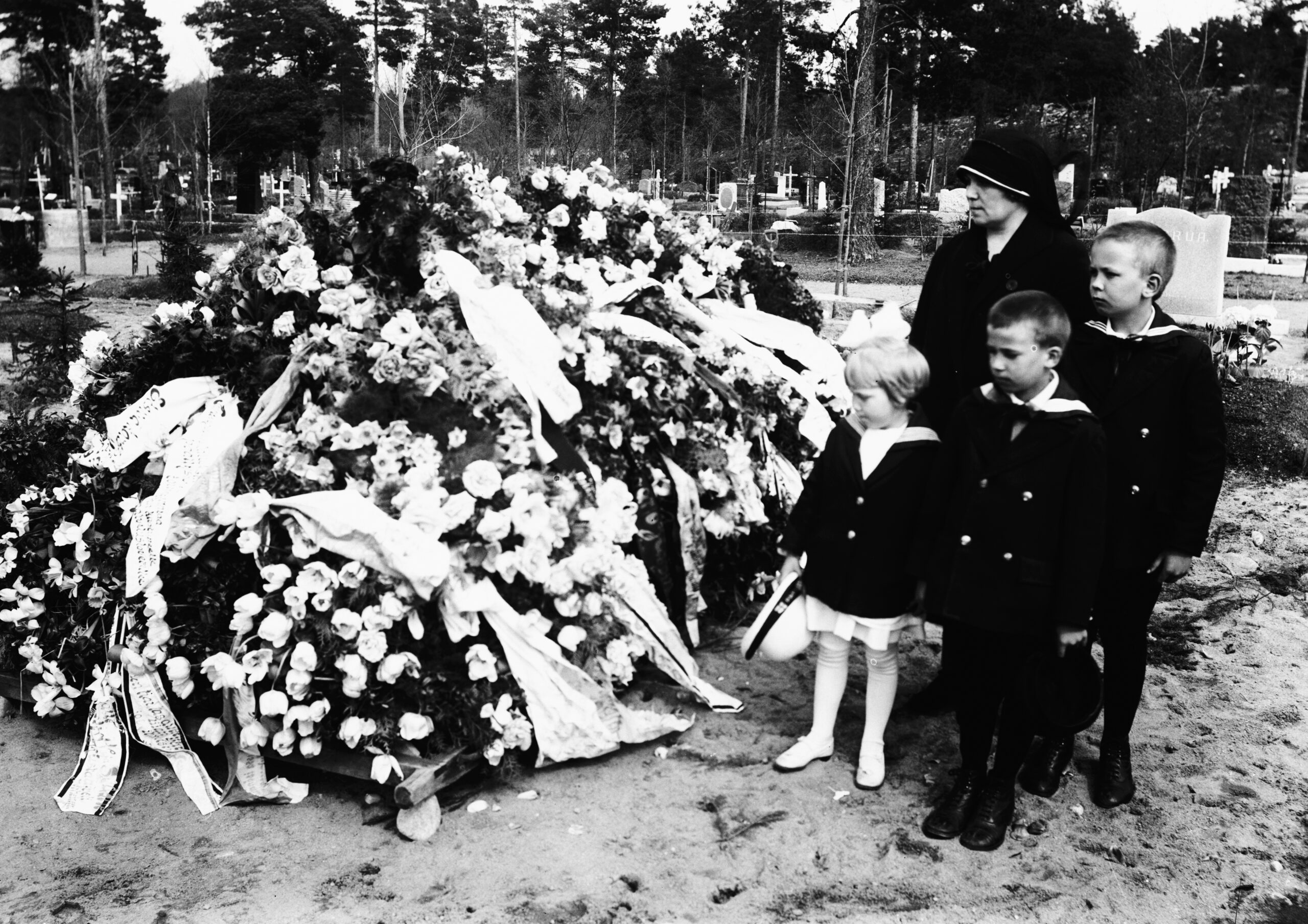 Mustavalkoinen kuva, jossa nainen ja kolme lasta seisovat lähekkäin haudalla, joka on peitetty seppeleillä ja kukilla.