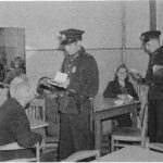 Mustavalkoinen kuva, jossa kaksi virkapukuista poliisia tutkii asiakirjoja pöytien ääressä istuvien henkilöiden lähellä. Oikealla on nainen ja vasemmalla mies.