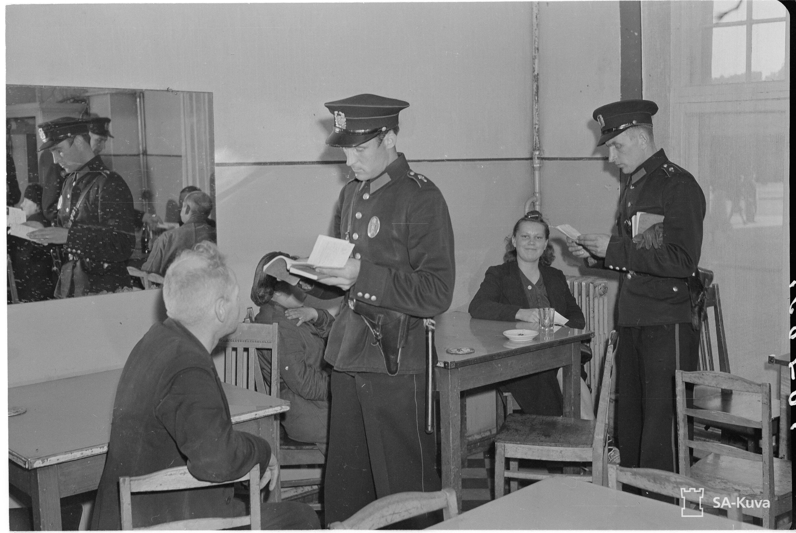 Mustavalkoinen kuva, jossa kaksi virkapukuista poliisia tutkii asiakirjoja pöytien ääressä istuvien henkilöiden lähellä. Oikealla on nainen ja vasemmalla mies.
