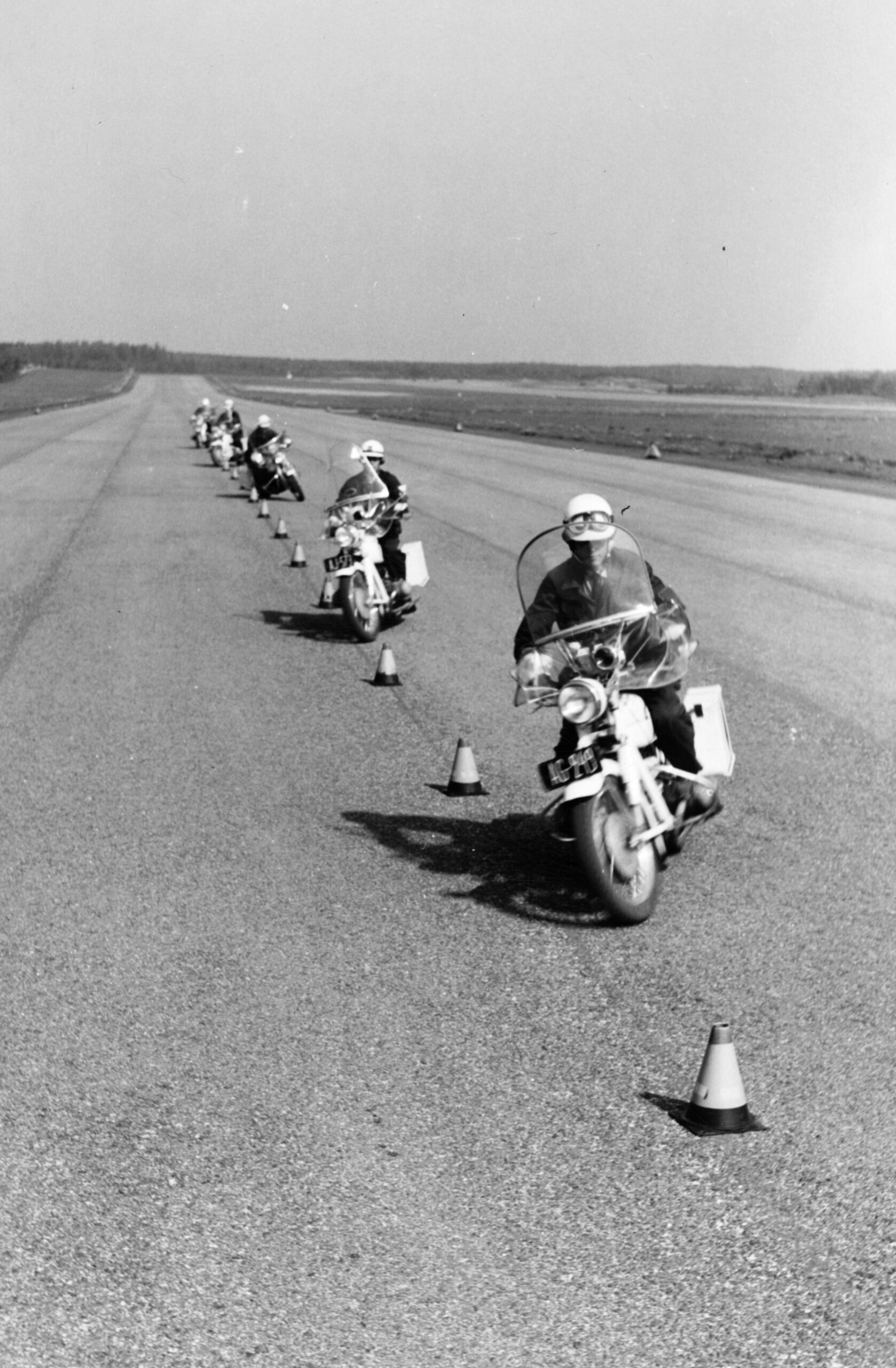Mustavalkoinen kuva harjoituksesta, jossa joukko moottoripyöräpoliiseja ajaa jonossa asfalttitiellä kartioiden lomassa. Poliiseilla on kypärät ja moottoripyörissä sivulaukut.