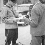 Mustavalkoinen kuva, jossa virkapukuinen poliisi keskustelee miehen kanssa ulkona. Molemmilla on paperia käsissään, ja miehen edessä maassa on matkalaukku. Taustalla näkyy auto.