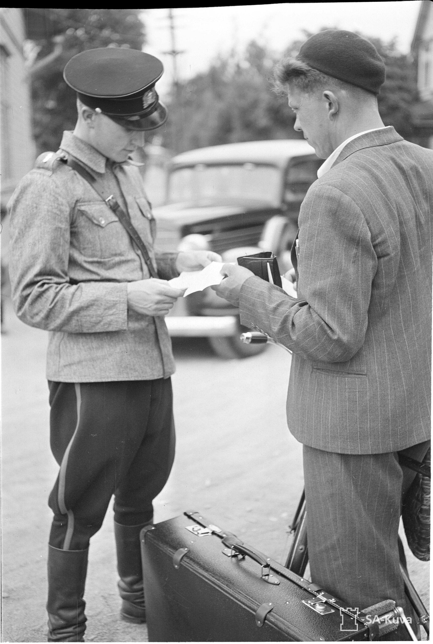 Mustavalkoinen kuva, jossa virkapukuinen poliisi keskustelee miehen kanssa ulkona. Molemmilla on paperia käsissään, ja miehen edessä maassa on matkalaukku. Taustalla näkyy auto.