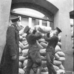 Mustavalkoinen kuva, jossa kaksi poliisia nostelee hiekkasäkkejä suojaukseksi rakennettuun pinoon ikkunan eteen. Vasemmalla seisoo kolmas poliisi tarkkailemassa työtä.