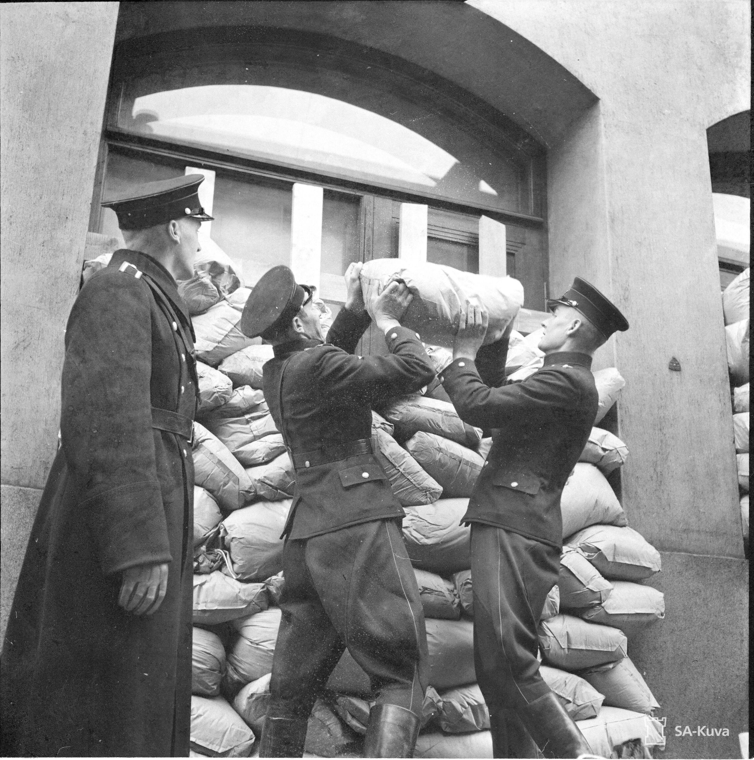 Mustavalkoinen kuva, jossa kaksi poliisia nostelee hiekkasäkkejä suojaukseksi rakennettuun pinoon ikkunan eteen. Vasemmalla seisoo kolmas poliisi tarkkailemassa työtä.