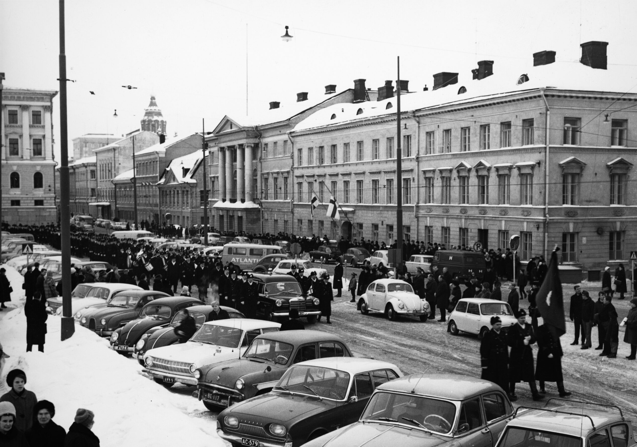 Mustavalkoinen kuva hautajaissaattueesta, jossa etummaisena auto ja auton ympärillä virkapukuisia poliiseja. Auton takana virkapukuisia poliiseja pitkässä jonossa. Osa kantaa soittimia. Kuva on talvinen. Taustalla näkyy suuria, klassisia rakennuksia, joiden katoilla on lunta.