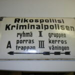 Värikuva vanhasta vaaleasta metallikyltistä, jossa on ohjeita ja tekstiä sekä suomeksi että ruotsiksi. Teksti kuuluu: "Rikospoliisi Kriminalpolisen", "ryhmä I gruppen", "porras A trappan", "kerros III våningen". Kyltissä on suuntaa osoittava nuoli.