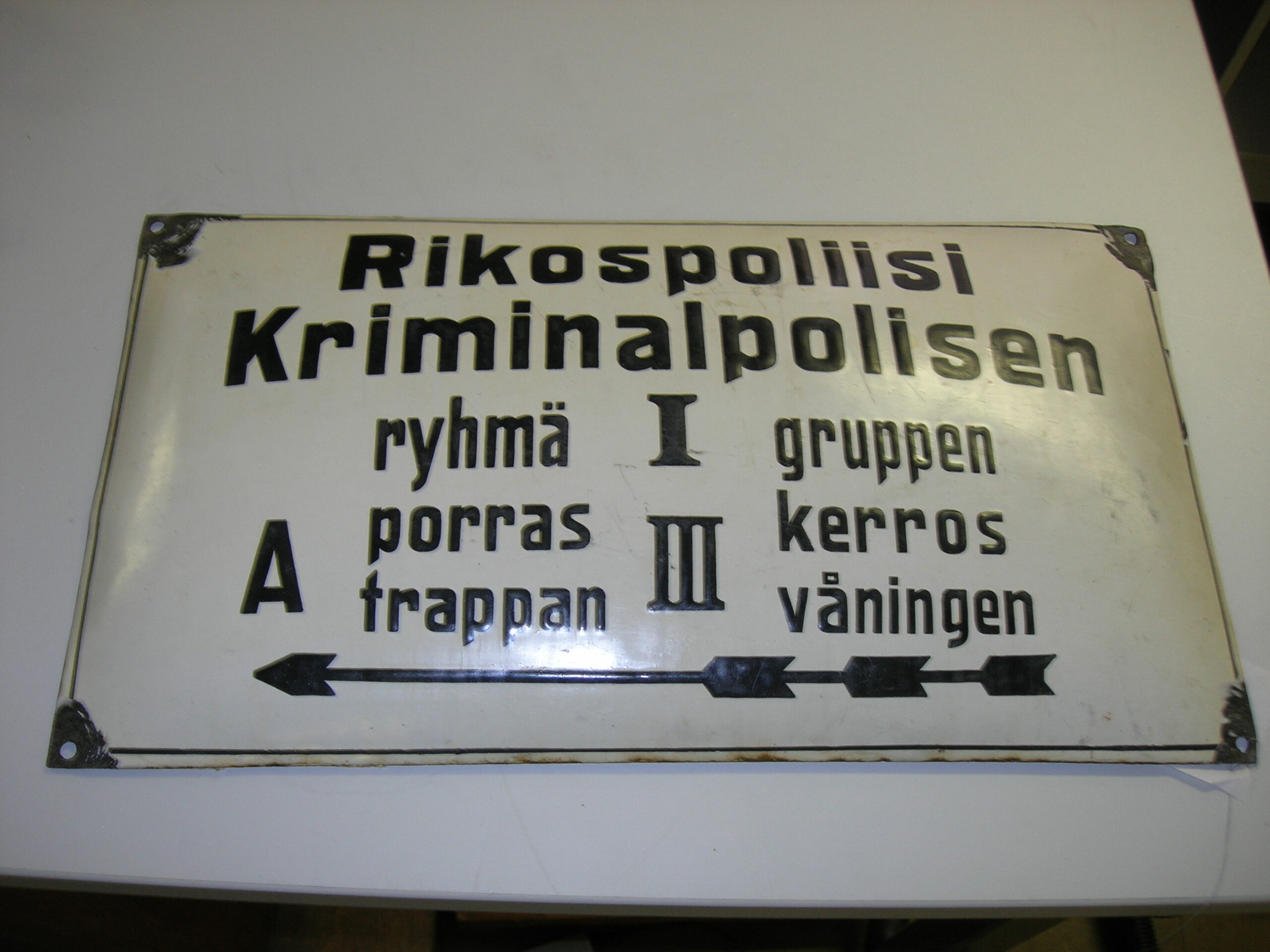 Värikuva vanhasta vaaleasta metallikyltistä, jossa on ohjeita ja tekstiä sekä suomeksi että ruotsiksi. Teksti kuuluu: "Rikospoliisi Kriminalpolisen", "ryhmä I gruppen", "porras A trappan", "kerros III våningen". Kyltissä on suuntaa osoittava nuoli.