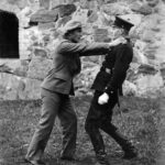 Mustavalkoinen kuva, jossa siviiliasuinen henkilö tarttuu käsillään virkapukuisen poliisin hartioihin. Taustalla näkyy kiviseinä.