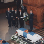 Värikuva kirkosta, jossa kaksi arkkua on peitetty Suomen lipuilla. Arkkuja ympäröi kukkia, ja ryhmä miehiä laskee seppeleitä muistotilaisuudessa.