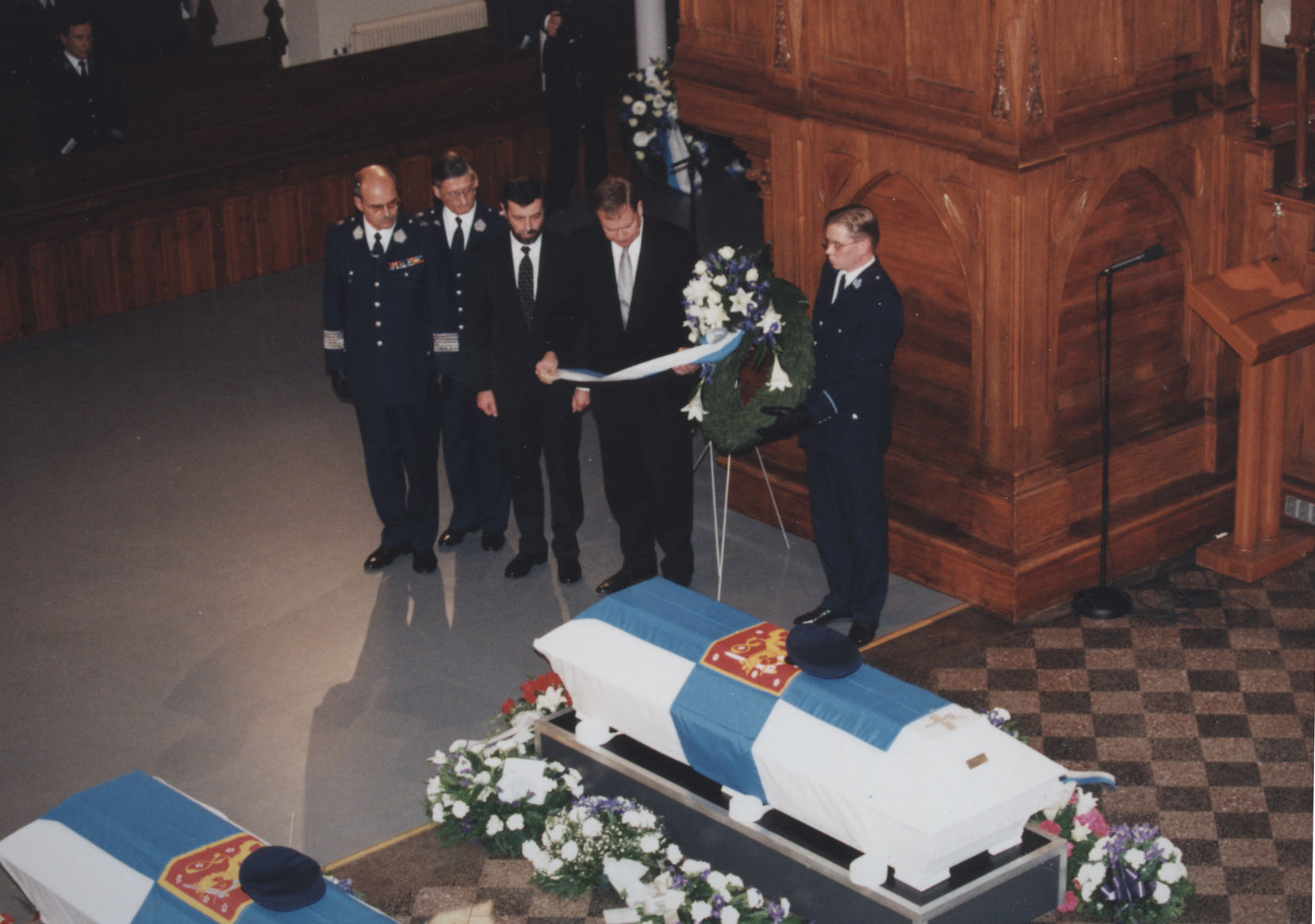 Värikuva kirkosta, jossa kaksi arkkua on peitetty Suomen lipuilla. Arkkuja ympäröi kukkia, ja ryhmä miehiä laskee seppeleitä muistotilaisuudessa.