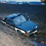 Värikuva Saab-poliisiautosta juuttuneena mutaiseen ojaan, kuljettajan puoleinen etuovi auki.