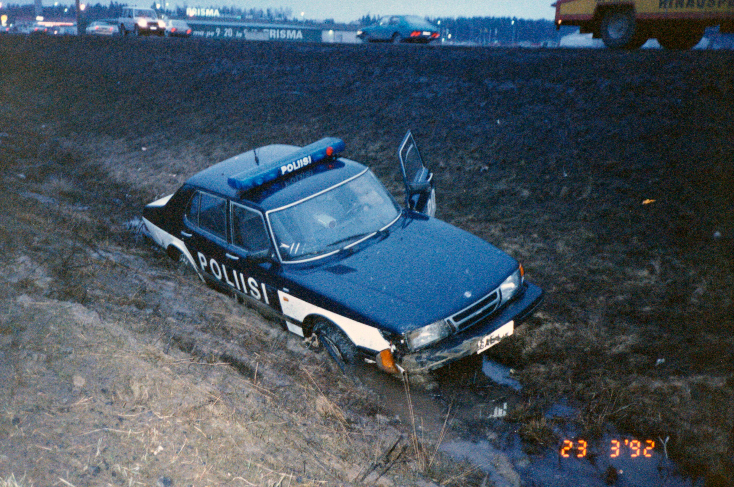 Värikuva Saab-poliisiautosta juuttuneena mutaiseen ojaan, kuljettajan puoleinen etuovi auki.