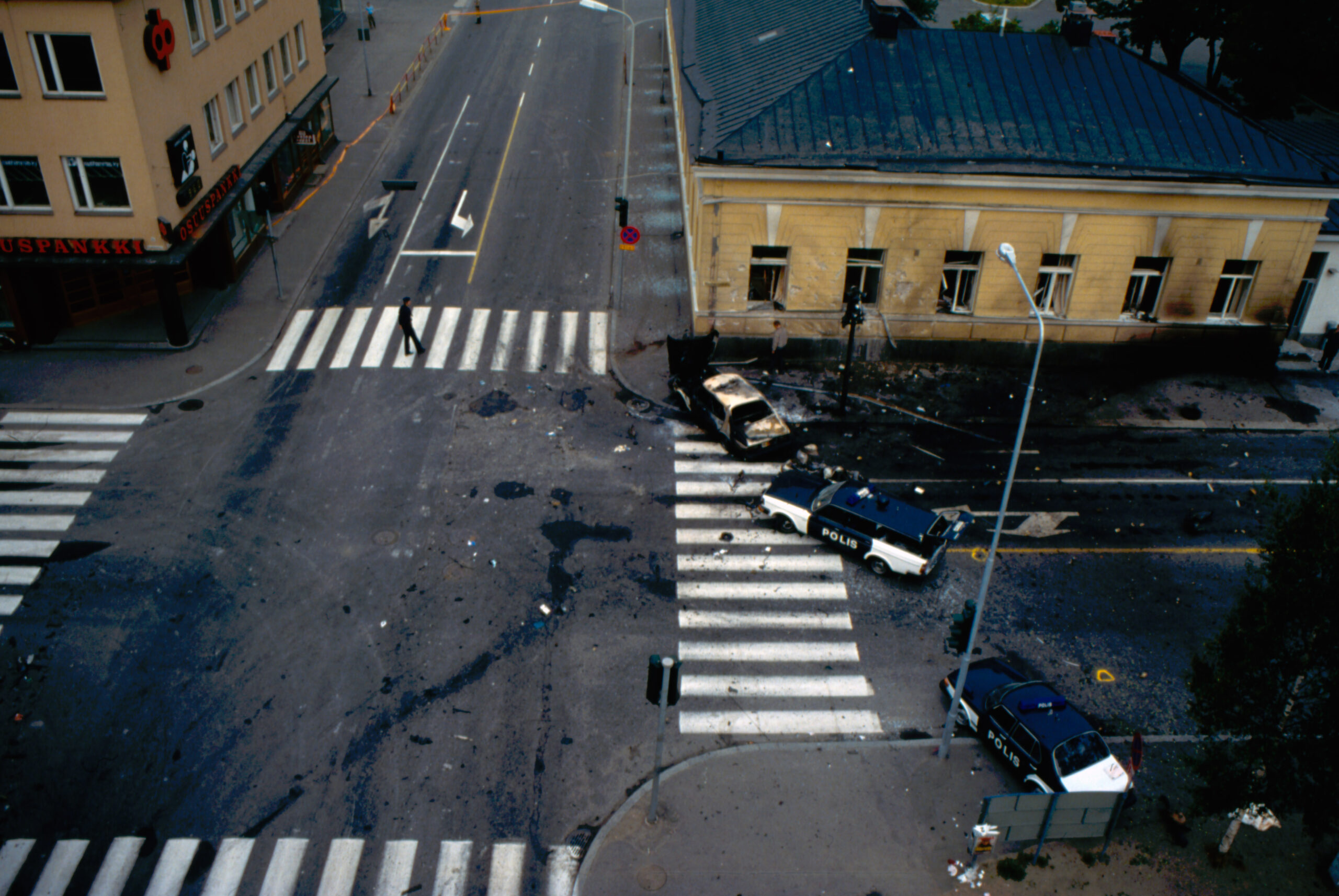 Ylhäältä otettu kuva kaupungin risteyksestä, jossa on suojateitä, yksi pahoin vahingoittunut vaalea henkilöauto ja kaksi poliisiautoa. Oikeapuoleisessa rakennuksia on myös vaurioita.