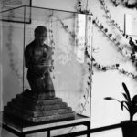 Kuvun sisällä veistos, jossa miesfiguuri on polvillaan portaiden päällä. Kädessä kypärä. Seinällä taulu, jossa tekstinä virkavala. Seinällä on köynnöskasvi.