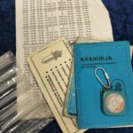 Värikuva esineistä: turkoosi ohut kirja, sen päällä avaimenperä, ja alla valkoisia papereita, joissa on luetteloita tai aikatauluja. Vasemmalla on kasa kapeita, kirkkaita putkia.