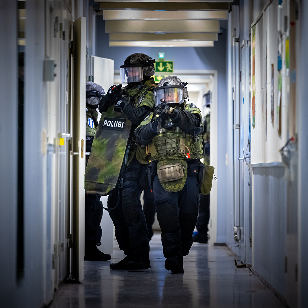 Värikuva, jossa raskaasti varustautuneita poliiseja liikkuu kapeassa käytävässä. Etualalla henkilö pitelee kilpeä, jossa lukee "POLIISI", ja kaksi muuta seuraa häntä. Käytävän seinillä on tauluja tai kuvia.