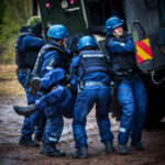 Värikuva, jossa ryhmä poliiseja raskaissa varusteissa on kantamassa henkilöä maastossa. Taustalla näkyy panssaroitu ajoneuvo.