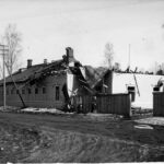 Mustavalkoinen kuvassa on vaurioituneita puutaloja kadun varrekka. Katon rakenteita ja peltiä on romahtanut. Vasemmalla näkyy vanha henkilöauto.