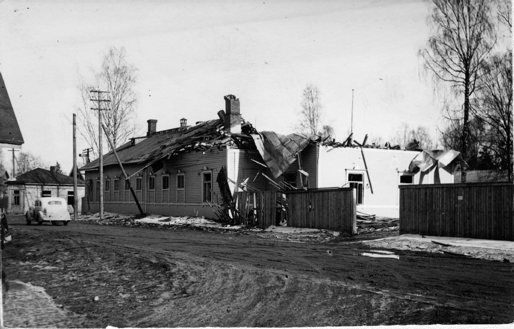 Mustavalkoinen kuvassa on vaurioituneita puutaloja kadun varrekka. Katon rakenteita ja peltiä on romahtanut. Vasemmalla näkyy vanha henkilöauto.