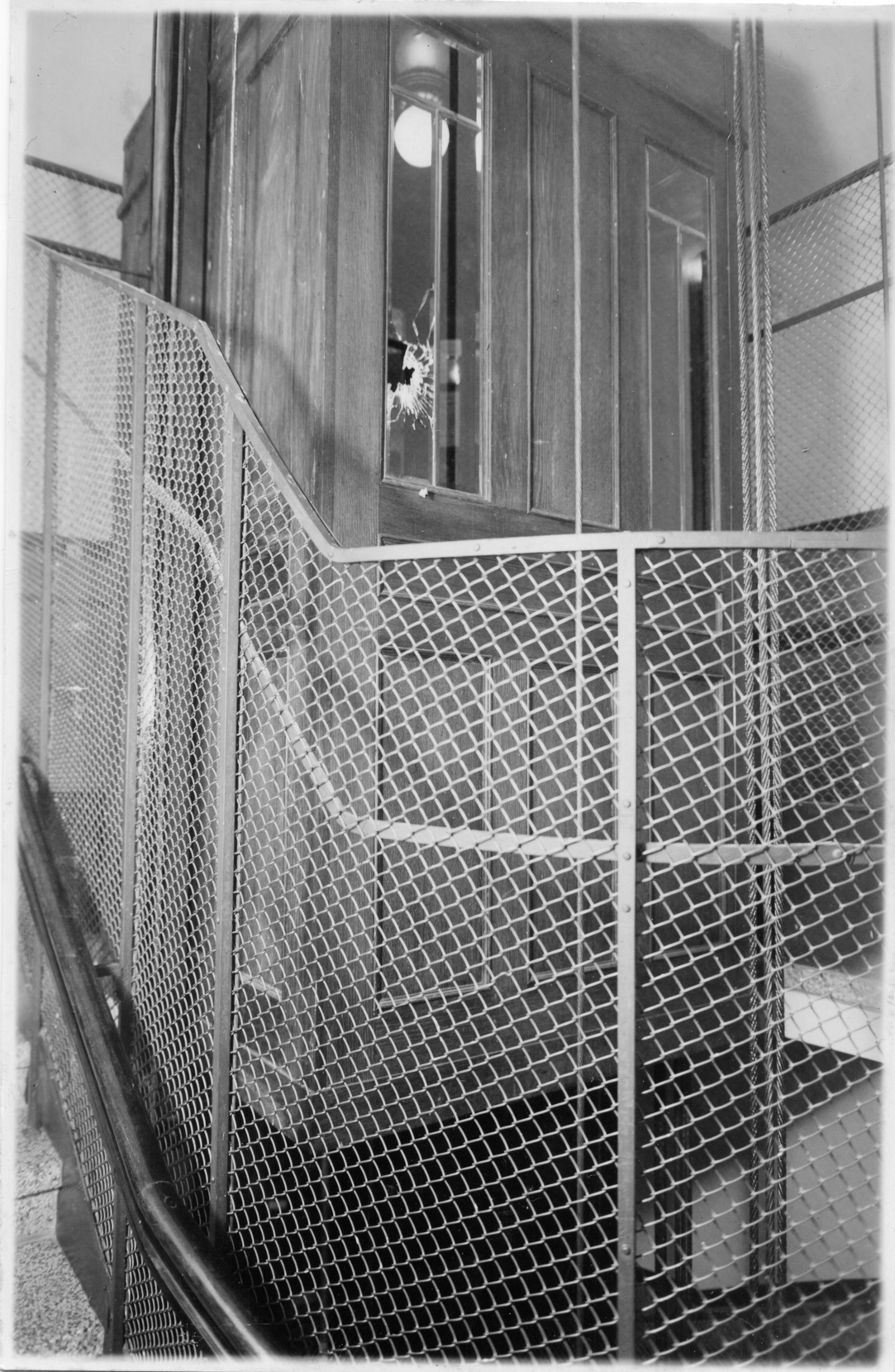 Mustavalkoinen kuva, jossa portaita kiertää metalliverkosta tehty kaide. Taustalla näkyy hissin seinä ja ovi, jonka ikkunassa on reikä.