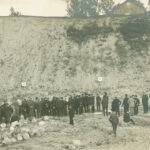 Mustavalkoinen ulkokuva, jossa suuri joukko ihmisiä on kokoontunut avoimelle alueelle hiekkapenkan eteen. Ihmiset seisovat ryhmissä, ja etualalla näkyy maassa palloja tai kiviä. Kauempana on vaaleita maalitauluja.