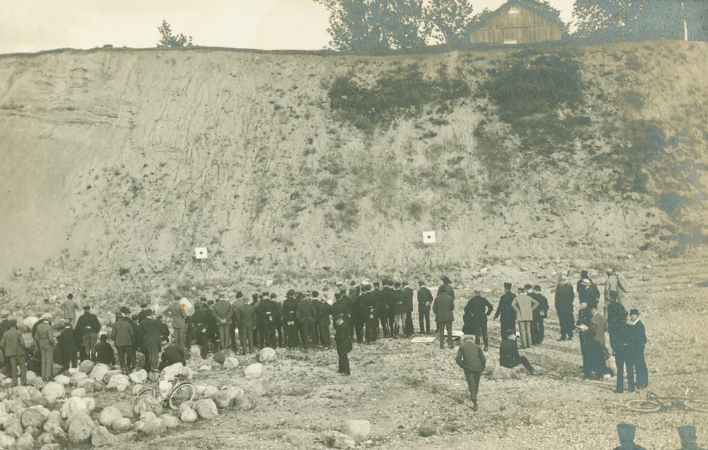 Mustavalkoinen ulkokuva, jossa suuri joukko ihmisiä on kokoontunut avoimelle alueelle hiekkapenkan eteen. Ihmiset seisovat ryhmissä, ja etualalla näkyy maassa palloja tai kiviä. Kauempana on vaaleita maalitauluja.