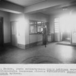 Mustavalkoinen sisäkuva: mies seisoo tiskillä tai luukulla valoisassa aulatilassa.