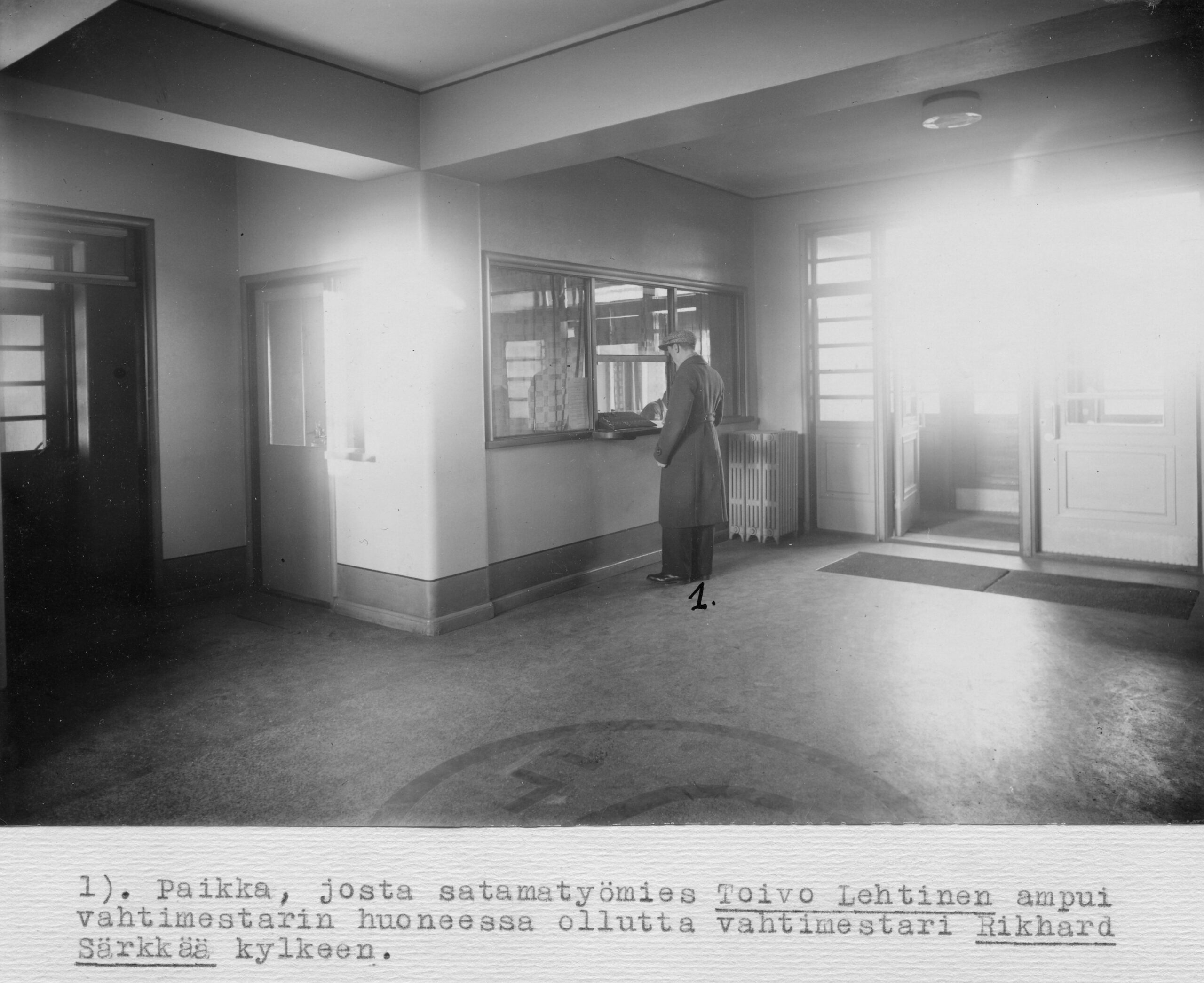 Mustavalkoinen sisäkuva: mies seisoo tiskillä tai luukulla valoisassa aulatilassa.