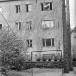 Mustavalkoinen kuva kolmikerroksisesta asuinrakennuksesta, jonka seinää peittää köynnöskasvi. Etualalla on hiekkaista maata, pensaita ja metallinen teline.