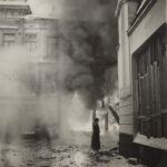 Mustavalkoisessa kuvassa katu on täynnä sankkaa savua. Keskellä seisoo tummiin pukeutunut poliisi, ja ympärillä näkyy kaupunkirakennuksia.