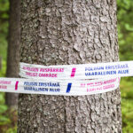 Värikuva, jossa poliisin käyttämää sulkunauhaa on kiedottu puunrungon ympärille metsässä. Nauhassa lukee "POLIISIN ERISTÄMÄ VAARALLINEN ALUE" ja ruotsiksi "AV POLISEN AVSÄRRT FARLIGT OMRÅDE".